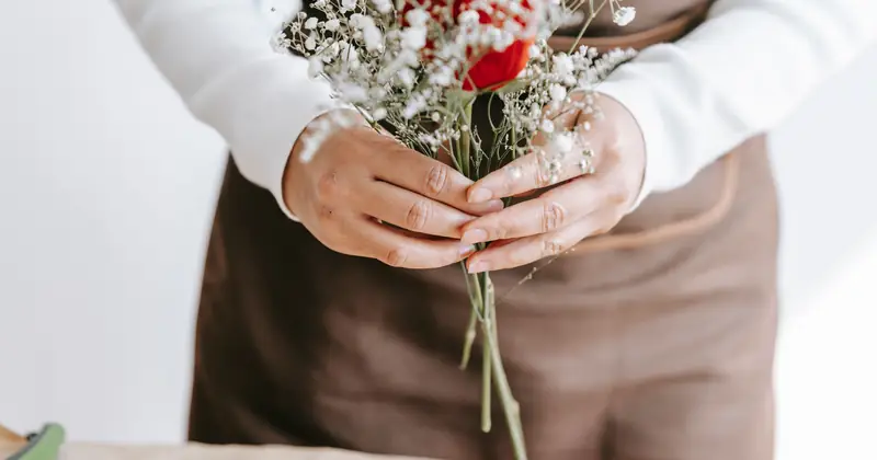 Crop unrecognizable woman in brown apron showing gentle bouquet with white flowers and bright red rose in blossom